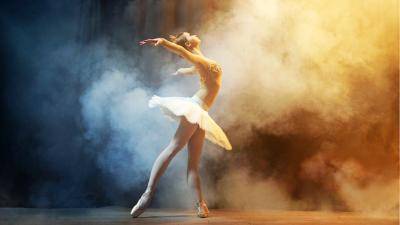 Ballerina in a white tutu posing sideways with leg extended in a tendu derrière position, slightly arched back with arms side, on a background of thick smoke.