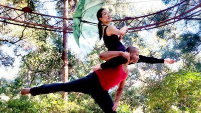 Outdoors surrounded by trees, male dancer in an arabesque lifting a female dancer on his shoulders as she is holding a green umbrella.