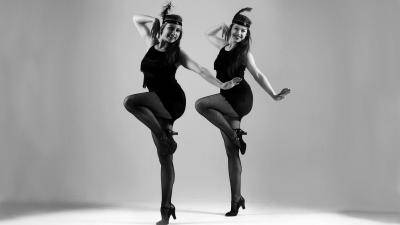 2 women dressed in 1920s flapper dancer outfits, doing a parallel passé.
