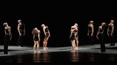 Group of dancers in black scattered across stage, standing upright, face to ceiling.