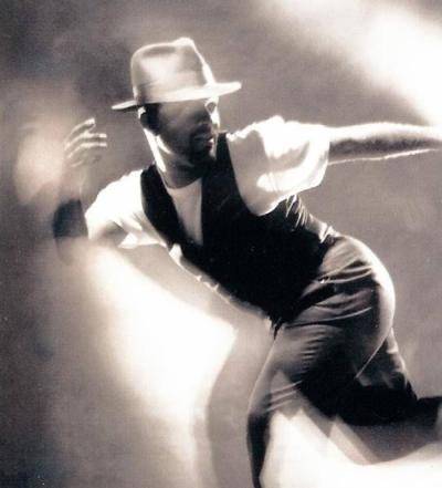 Dance educator Carlos Jones wearing a white t-shirt, black vest and a hat, in a dance move looking back