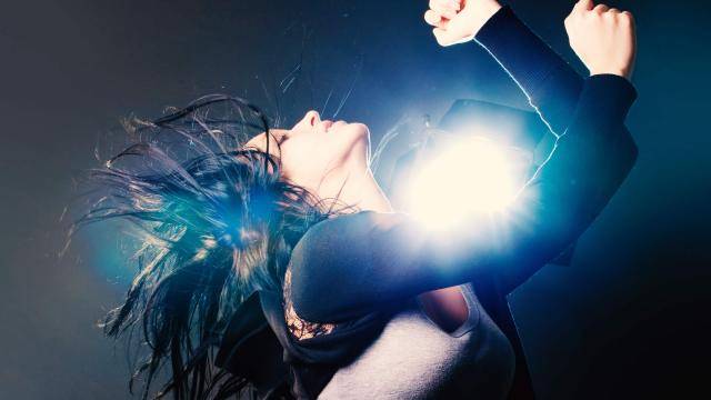Girl throwing her black hair back, bent arms up with fists, in front of a bright set light