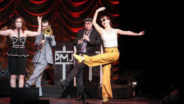 Demi Remick in yellow pants, kicking one leg up with the Postmodern Jukebox band in the background