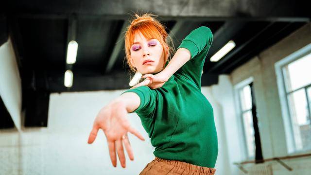 Redhead woman with a green top, reaching out with one hand, in dance studio.