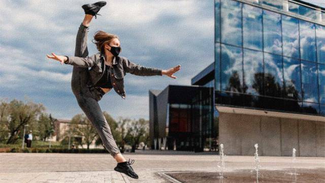 dancer wearing a mask in a split jump, in front of a glass building