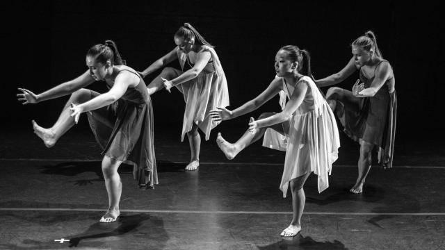 4 young female dancers on stages, with dresses, contracting over their lifted leg.