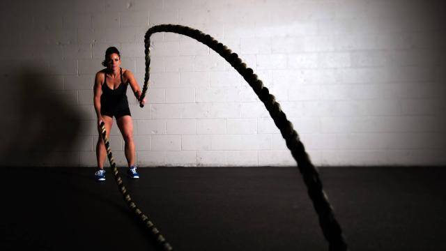 Woman in black short and tank top in front of a white wall, working out with big ropes.