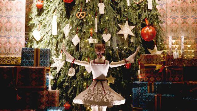 A puppet ballerina in a white tutu with red trim raising her arms towards a tall decorated christmas tree she is facing.