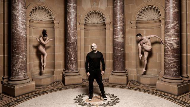 Rafael Bonachela standing in the middle of a greek alcove at the National Gallery of Australia, with 2 nude dancers on either side posing as statues.